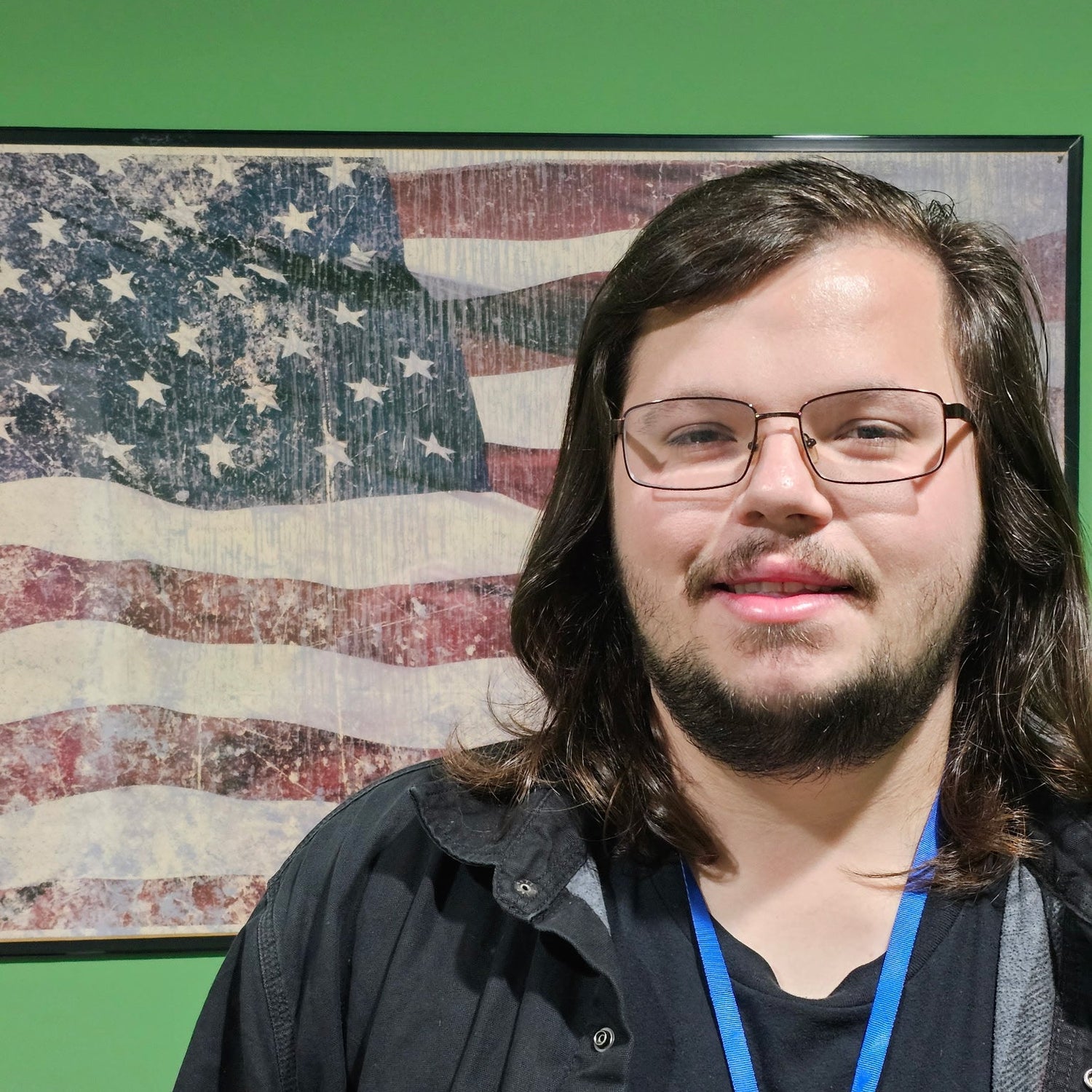 Person with glasses and a beard standing in front of a framed American flag on a green wall.